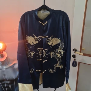 Mörkblå sidenjacka med drakbroderi - Säljer en elegant mörkblå sidenjacka från TianBaoTong med detaljerade drakbroderier i guld på framsidan. Jackan har en traditionell design med knappar och långa ärmar. Perfekt för en unik och stilfull look.