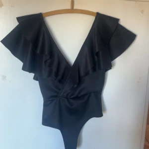 Svart body från Gina Tricot - Elegant svart body från Gina Tricot med volangdetaljer och v-ringad design och öppen rygg. Nyskick, storlek S❤️