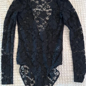 Svart spetsbody - Snygg svart spetsbody med långärmad design och öppen v-ringning därbak. Perfekt för en elegant look. Knäppning i grenen för enkel användning.