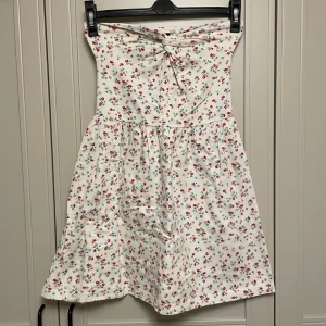 Blommig klänning från H&M Divided - Charmig axelbandslös klänning från H&M Divided med ett blommigt mönster i rött och blått på en vit bakgrund. Klänningen har en knytdetalj framtill och en avslappnad passform som passar perfekt för varma dagar.
