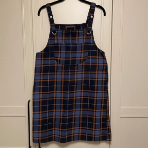 Rutig jeansklänning från Monki - Säljer en cool rutig klänning från Monki i blått och orange. Klänningen har justerbara axelband med knappar och en praktisk ficka framtill. Perfekt för en avslappnad stil! I ett tunnare jeans material.