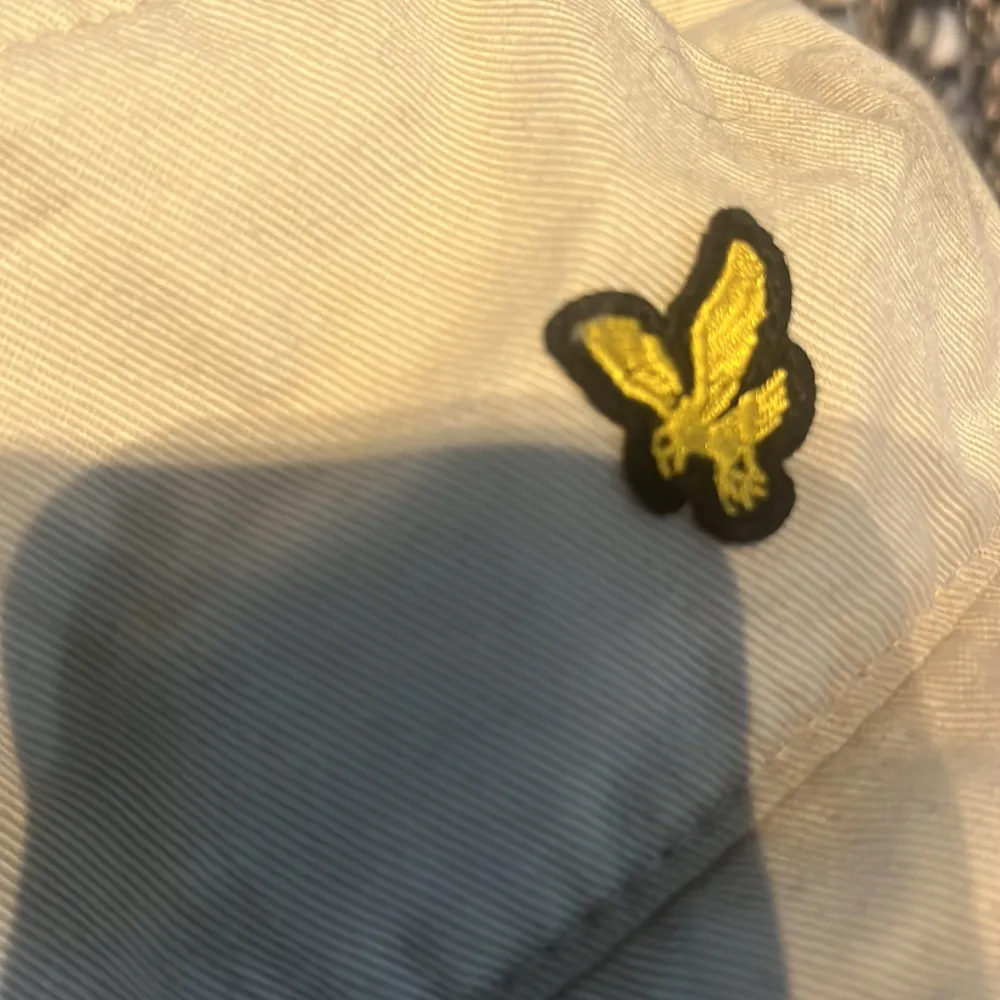 Snygg beige bucket hat från Lyle & Scott med deras ikoniska gula logga framtill. Perfekt för en avslappnad stil och skydd mot solen. Tillverkad i bomull för en bekväm passform.. Asusteet.
