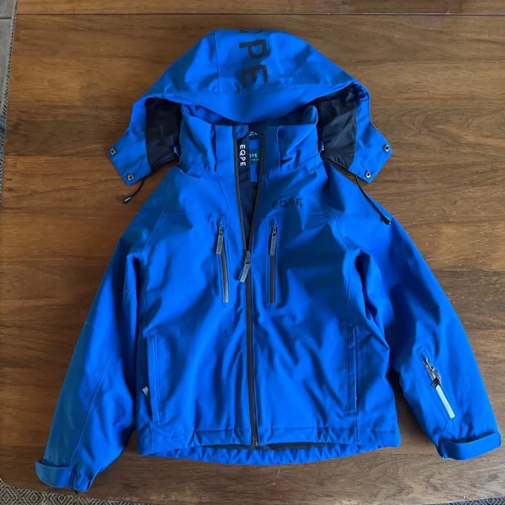 Snygg blå vindjacka från Gore med justerbar huva och flera praktiska fickor med dragkedja. Perfekt för blåsiga dagar och utomhusaktiviteter. Jackan har långa ärmar och en stilren design.. Takit.
