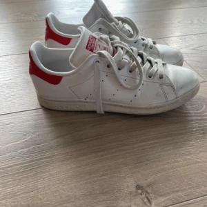 Vita Adidas Stan Smith med röda detaljer - Säljer ett par klassiska vita Adidas Stan Smith sneakers med röda detaljer.