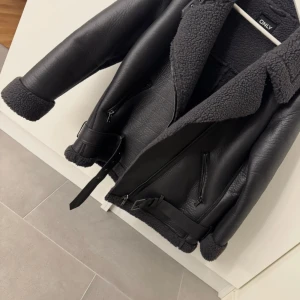 Svart jacka från ONLY - Snygg svart jacka från ONLY med fuskpälsfoder och krage. Jackan har dragkedja och bälte i midjan för en cool look. Använt en gång.