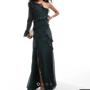 Mörkgrön oneshoulder balklänning/finklänning - Elegant mörkgrön oneshoulder klänning i nyskick (enbart provad, lappen kvar) med volangdetaljer och slits. Perfekt för bal, finmiddag eller bröllop! Den har en lång ärm och en asymmetrisk design som ger en unik look. Köpt för 929kr på ASOS.