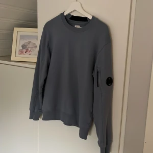 Grå sweatshirt från C.P. Company - Säljer en stilren grå sweatshirt från C.P. Company. Tröjan har långa ärmar och en rund halsringning. På ärmen finns en snygg detalj med en liten ficka och en rund logotyp. Perfekt för en avslappnad stil.