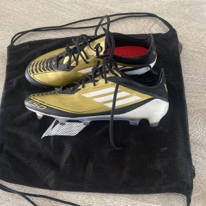 Guld och svarta fotbollsskor från Adidas - Säljer ett par snygga fotbollsskor från Adidas i guld och svart med vita detaljer. Skorna har snörning och en stilren design med tre vita ränder på sidan. Perfekta för fotbollsplanen! Dom är använda ett fåtal gånger men annars har de inga skador.