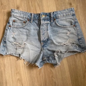 Ljusa jeansshorts med slitningar - Säljer ett par ljusa jeansshorts med coola slitningar och fransiga kanter. De har en klassisk femficksdesign och knappgylf. Perfekta för en avslappnad sommarlook!