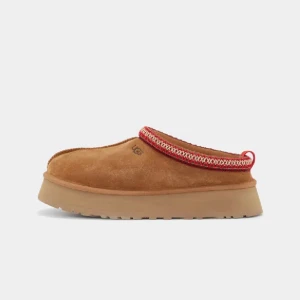 Bruna mocka tofflor från UGG - Snygga bruna tofflor i mocka från UGG med en dekorativ röd kant. Perfekta för att hålla fötterna varma och bekväma. Den robusta sulan ger bra grepp och komfort. Slutsåld. Använda 5 ggr kanske
