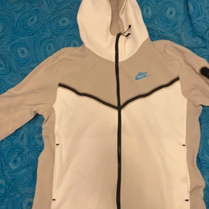 Vit grå tracksuit från Nike - Säljer en vit grå tracksuit från Nike med en blå logga på sidan. Byxorna har en elastisk midja med snörning och är perfekta för en avslappnad stil. Pris kan diskuteras. Sparsamt använd