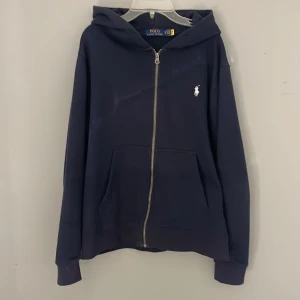 Mörkblå hoodie från Polo Ralph Lauren - Säljer en stilren mörkblå hoodie från Polo Ralph Lauren med dragkedja med ett 6/10 skick den har mäst bara legat i garderoben därflr säljare jag den är storlek M men känd som en större S i storleken 
