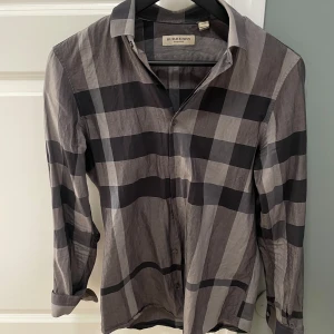 Rutig skjorta från Burberry - Snygg rutig skjorta från Burberry i grå och svart. Skjortan har långa ärmar och klassisk krage med knappar framtill. Perfekt för en stilren look.
