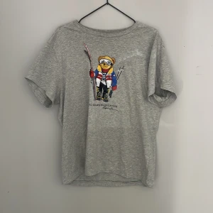 Grå t-shirt med Polo Bear från Ralph Lauren - Säljer en grå t-shirt från Ralph Lauren med en cool Polo Bear-tryck på magen tröjan är i ett topp skick.  Passar som en vanlig M i storleken 