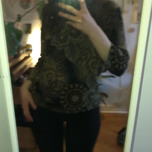 Mönstrad tröja från Tradition - Säljer en unik mönstrad tröja från Tradition i svart och guld. Tröjan har ett intrikat mönster med blommor och spiraler, och är tillverkad i polyester. Perfekt för att ge din outfit en extra touch!