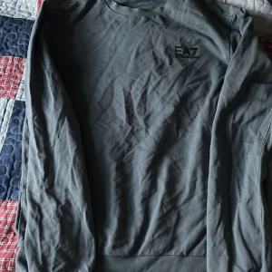 Grå tröja från Emporio Armani EA7 - Säljer en stilren grå tröja från Emporio Armani EA7. Tröjan har en enkel design med långärmade ärmar och en rund halsringning. Perfekt för en avslappnad och trendig look.