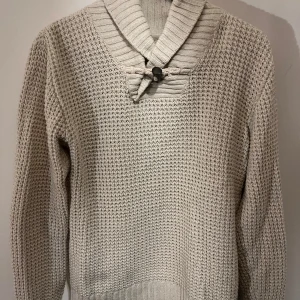 Beige stickad tröja med krage - Säljer en stilren beige stickad tröja med en unik krage och knappdetalj framtill. Tröjan har långa ärmar och ett texturerat mönster som ger en mysig känsla. Perfekt för kyligare dagar och sommarkvällar. 158-164 är mer som en small!!!