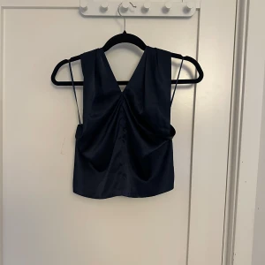 Svart ärmlös topp - Snygg svart ärmlös topp med djup v-ringning och draperad design. Perfekt för en stilren och elegant look. Tillverkad i ett glansigt material som ger en lyxig känsla.