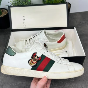 Vita sneakers med broderad detalj från Gucci - Snygga vita sneakers från Gucci med broderad hunddetalj på sidan. Skorna har klassiska gröna och röda ränder samt snörning. Perfekta för en stilren look.