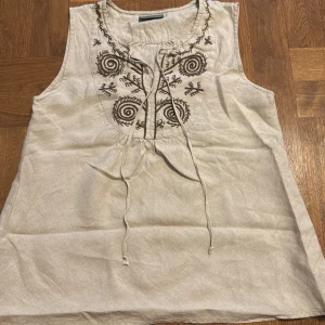Snygg blus - Säljer en stilren beige ärmlös topp i 100% linne från Nygårdsanna. Toppen har ett vackert broderat mönster i brunt framtill och knytband vid halsen. Perfekt för varma sommardagar!