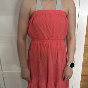 Korallfärgad axelbandslös klänning - Snygg korallfärgad axelbandslös klänning med elastisk midja och volangdetaljer längst ner. Perfekt för varma sommardagar.