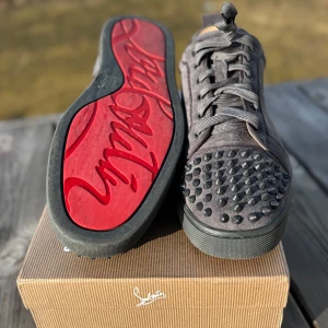 Louboutins  - Snygga grå sneakers i mocka från Christian Louboutin med coola nitar på tån och sidorna. Skorna har en klassisk röd sula och snörning framtill. Perfekta för att ge din outfit en edgy touch.