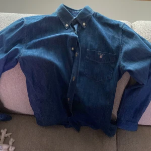 Blå jeansskjorta från GANT - Säljer en snygg blå jeansskjorta från GANT med klassisk krage och knappar framtill. Skjortan har en bröstficka med GANT-logga och långa ärmar. Perfekt för en avslappnad stil.