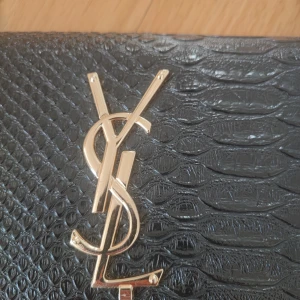 Svart axelväska från YSL - Snygg svart axelväska från YSL med en elegant guldlogga på framsidan. Väskan har en texturerad yta som påminner om reptilmönster och en stilren guldkedja som axelrem. Perfekt för att ge en lyxig touch till din outfit.
