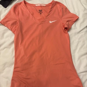 Korallfärgad t-shirt från Nike Pro - Säljer en snygg korallfärgad t-shirt från Nike Pro med v-ringning och korta ärmar. Perfekt för träning eller en aktiv vardag. Den har en liten vit Nike-logga på bröstet. Materialet är lätt och andas bra.