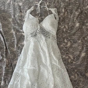 Vit spetsklänning med knytband - Charmig vit klänning med spetsdetaljer och knytband i nacken. Klänningen har en v-ringad design och öppen rygg, vilket ger en luftig och somrig känsla. Perfekt för varma dagar!