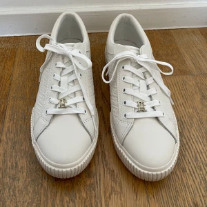 Vita sneakers från Tommy Hilfiger strl 40 - Snygga vita sneakers från Tommy Hilfiger med snörning och ett diskret mönster på sidorna. Skorna har  en stilren design med märkets logotyp på sulan. Strl 40. 