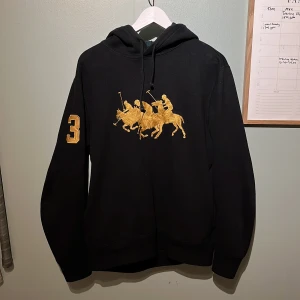 Svart hoodie från Polo Ralph Lauren - Säljer en svart hoodie från Polo Ralph Lauren med en snygg luva med flanellmönster samt unik och stilig detalj på framsida och ena ärmen! Tröjan är sparsamt använd och i fint skick😁 Hör av dig vid funderingar eller prisförslag😉🤝
