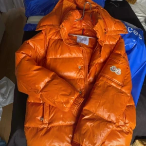 Orange dunjacka från Moncler - Säljer en snygg orange dunjacka från Moncler (at kod finns i jackan) med en quiltad design och dragkedja. Jackan har långa ärmar och en praktisk huva. Perfekt för kyliga dagar och ger en färgklick till din outfit. Priset kan diskuteras något då det inte är riktigt säsong just nu och den behöver lämna plats i min garderob.