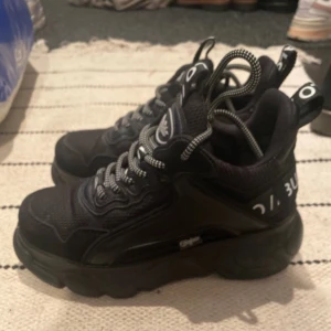 Svarta sneakers från Buffalo - Snygga svarta sneakers från Buffalo med tjock sula och coola detaljer. Skorna har snörning och märkets logga syns tydligt på både plösen och baksidan. Perfekta för en trendig look.