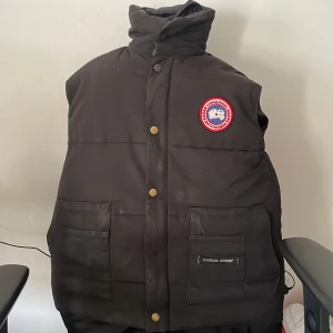 Svart dunväst från Canada Goose - Säljer en svart dunväst från Canada Goose med hög krage och knappar framtill. Västen har två stora fickor och en broderad logotyp på bröstet. Perfekt för kyliga dagar!