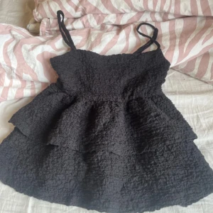 Svart volangtopp från Gina Tricot - Säljer en svart topp från Gina Tricot med smala axelband och en texturerad yta. Toppen  har en volangdetalj som ger en fin form och är perfekt för olika tillfällen. Passar bra för dig som gillar en stilren look. 