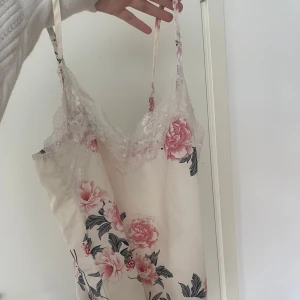 Blommigt linne med spetsdetaljer - Säljer ett linne med blommigt mönster i rosa och grått på en vit bakgrund. Klänningen har spetsdetaljer vid halsringningen och smala axelband. Perfekt för sommarens varma dagar. Använd men i bra skick, strl är obefintlig men jag har storlek S, skulle säga att den passar S-L