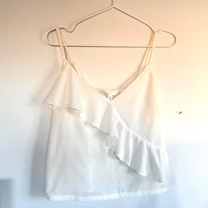 Vit topp med volang - Säljer en stilren vit topp med smala axelband och en snygg volangdetalj framtill. 