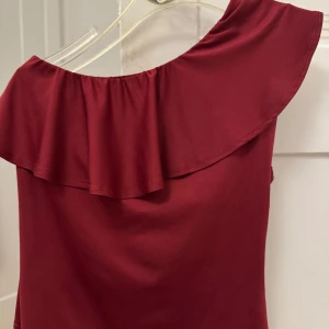 Vinröd enaxl topp med volangdetalj - Snygg vinröd topp med en elegant volangdetalj vid halsen. Perfekt för en stilfull kväll. Toppen har en mjuk och följsam passform. 