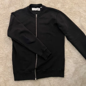 Svart tröja från L'Homme Rouge - Säljer en stilren svart tröja från L'Homme Rouge i 100% bomull. Tröjan har en perfekt minimalistisk look. Skicket är som nytt förutom två små skrapningar vid baksidan av armbågen. 