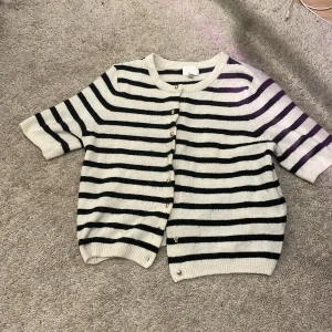 Randig kofta från H&M - Säljer en stilren randig kofta från H&M i svart och vit. Koftan har korta ärmar och knäpps med eleganta guldfärgade knappar. Perfekt för en avslappnad look.