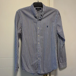 Randig skjorta från Ralph Lauren - Säljer en stilren blå- och vitrandig skjorta från Ralph Lauren i slim fit. Skjortan har långa ärmar och en klassisk krage med knappar framtill. Perfekt för en elegant look.