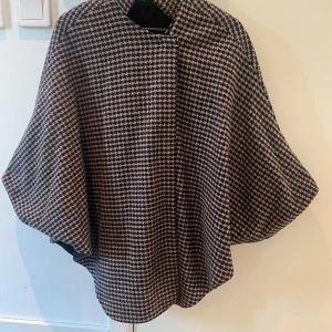 Rutig poncho kappa - Snygg rutig kappa i svart och beige med en klassisk design. Perfekt för att hålla stilen under vårens dagar. Den har en lös passform och är ett tidlöst tillskott till garderoben. Köpte den i Paris på en marknad, av en dam som sydde.