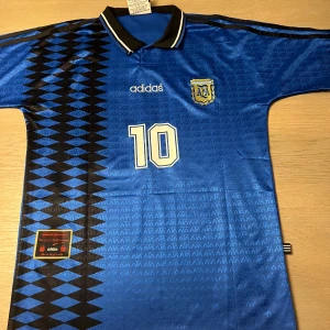 Blå fotbollströja från Adidas - Säljer en blå fotbollströja från Adidas med nummer 10 och texten 'Maradona' på ryggen. Tröjan har ett svart mönster längs sidan och en vit Adidas-logga på framsidan. Perfekt för fotbollsfans!
