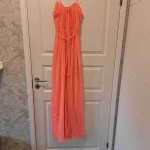 Persikofärgad långklänning från Vila - Säljer en vacker persikofärgad långklänning från Vila. Klänningen har smala axelband och en knytning i midjan för en fin passform. Perfekt för sommarens festligheter!
