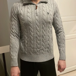 Grå stickad tröja från Polo Ralph Lauren - Säljer en stilren grå stickad tröja från Polo Ralph Lauren med kabelstickat mönster och dragkedja vid halsen. Tröjan har långa ärmar och en broderad logotyp på bröstet. Perfekt för kyligare dagar.