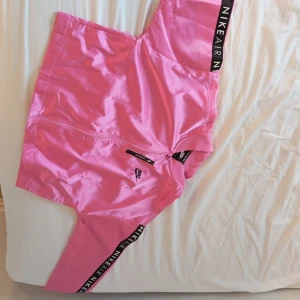 Rosa jacka från Nike - Snygga rosa shorts från Nike med svart resårband där det står 'Nike Air'. Perfekta för en sportig look eller avslappnad stil. De har en dragkedja framtill och är gjorda i ett glansigt material.