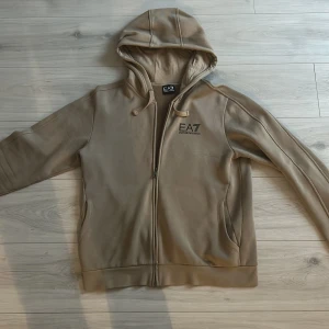 Beige hoodie från Emporio Armani - Säljer en stilren beige hoodie från Emporio Armani med dragkedja och huva. Tröjan har långa ärmar och EA7-logga på bröstet. Perfekt för en avslappnad stil.