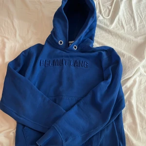 Blå hoodie från Helmut Lang - Säljer en snygg blå hoodie från Helmut Lang med broderad logga på bröstet. Tröjan har en klassisk känguruficka och justerbar huva med dragsko. Perfekt för en avslappnad stil.