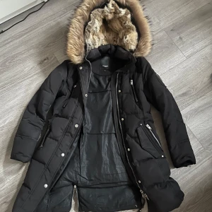 Svart dunjacka från Rockandblue - Säljer en svart dunjacka från Rockandblue med pälskantad huva. Jackan har dragkedja och knappar framtill samt flera praktiska fickor. Perfekt för kalla dagar med sin långa design och vadderade stil. Äkta päls men dock ett hål i höger armhåla.